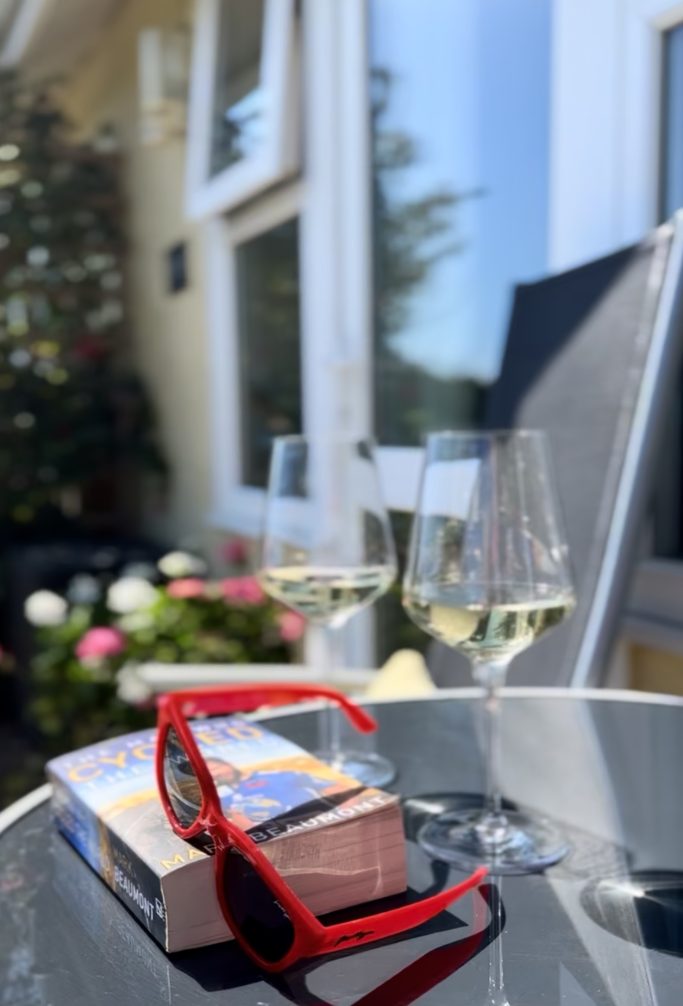 Two glasses of wine, a book, and sunglasses on the sunlit patio table outside 19 Evening Star chalet, Isle of Wight.